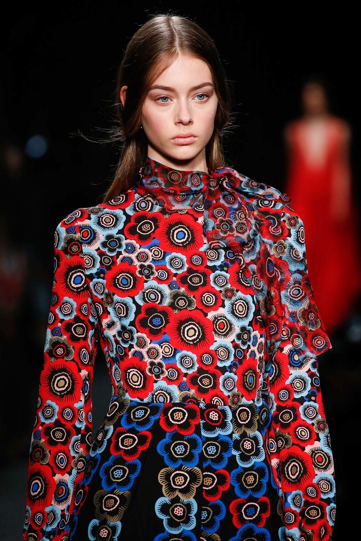 a young woman model is showcased on a runway, wearing a striking longsleeved dress. the dress is predominantly black, adorned with a vibrant floral print featuring red, blue, and beige flowers. the print's style is bold and eyecatching.  the model has light brown hair styled simply, with minimal makeup enhancing her features. the background is blurry, indicating a fashion show setting, with another model partially visible in a red dress in the background. the overall image focuses on the model and her dress, highlighting the intricate design and colors of the garment.