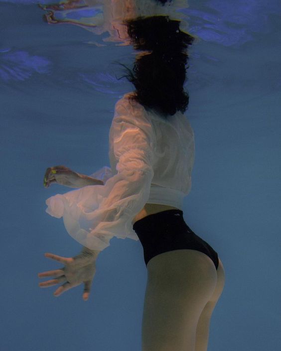 a woman wearing a white blouse and black bikini bottoms is submerged in a pool of blue water. her hair is floating above her head and her arms are outstretched. the perspective is from underwater, looking up towards the surface. the lighting is natural and creates a dreamy atmosphere.