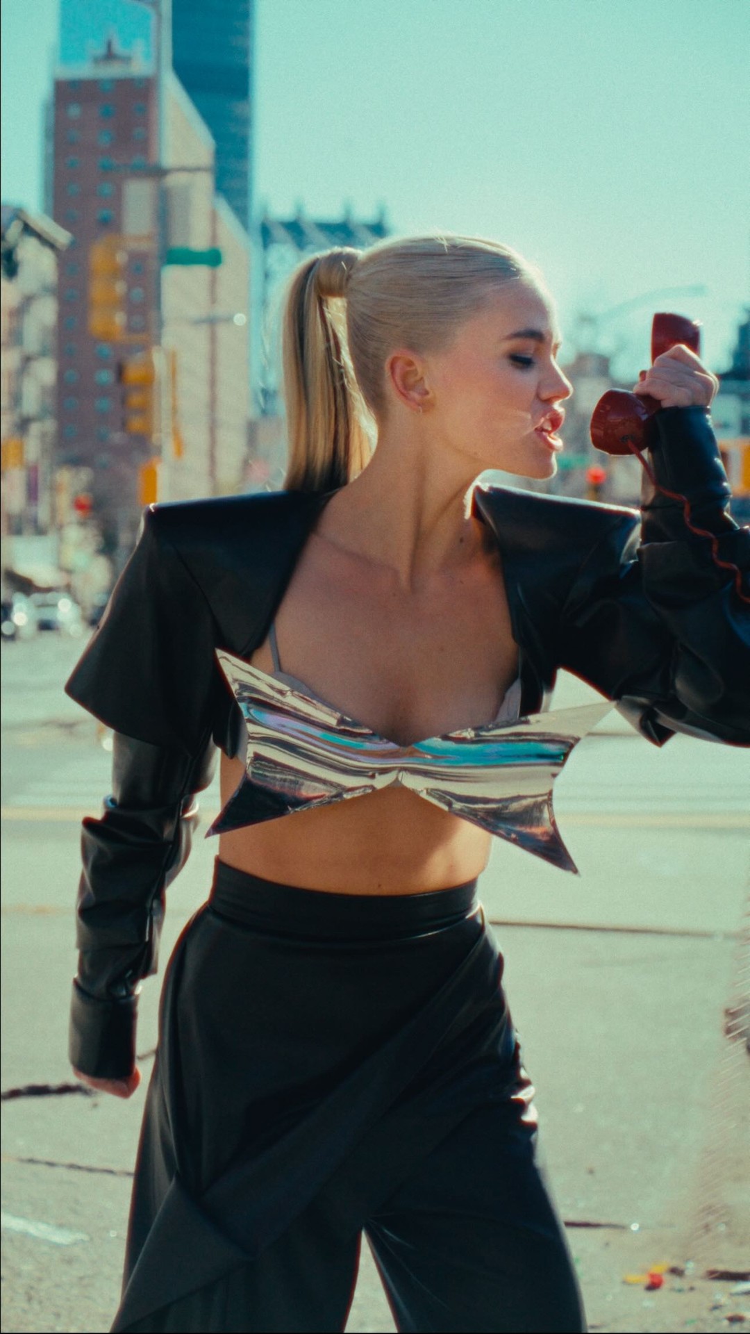 a woman with blonde hair in a ponytail is standing on a street with a building in the background. she is wearing a black jacket, a silver and blue top, and black pants. she is holding a red telephone to her mouth.