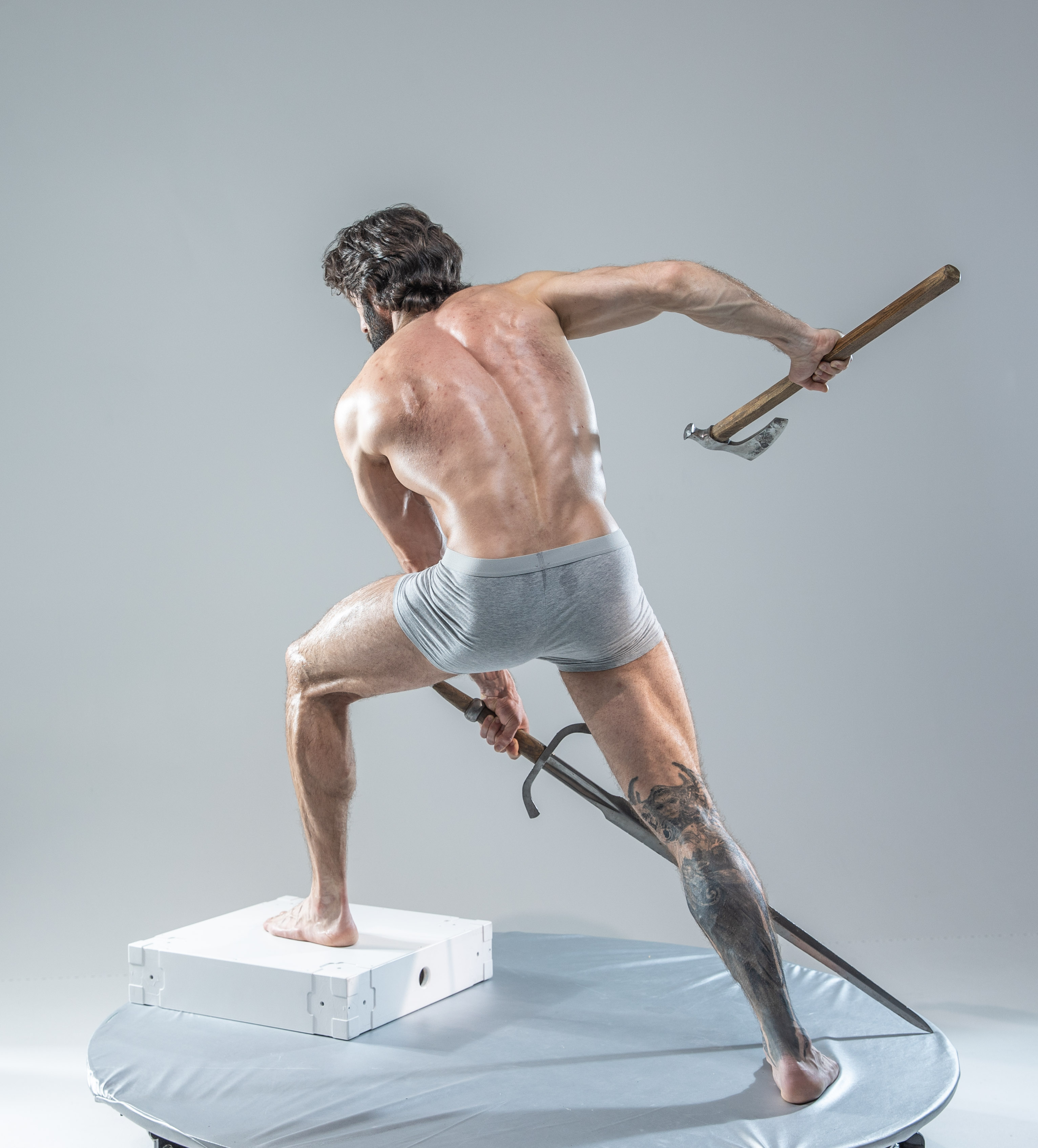 a muscular man with long hair and a beard stands in a dynamic pose on a round platform, his back to the camera. he is dressed only in gray underwear, showcasing his physique.  he holds a sword in his right hand, pointed downwards, and an axe in his left, raised above his head. the image appears to be a studio shot with controlled lighting, highlighting the man's muscles and the weapons he holds.