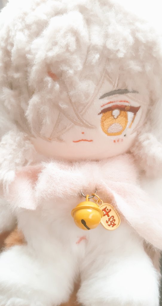 this is a close up photo of a white fluffy plushie that resembles an anime or cartoon character. it is wearing a pink scarf and has a yellow bell attached to it. it's large eyes are yellow, and it has a red mouth. the plushie is very cute.
