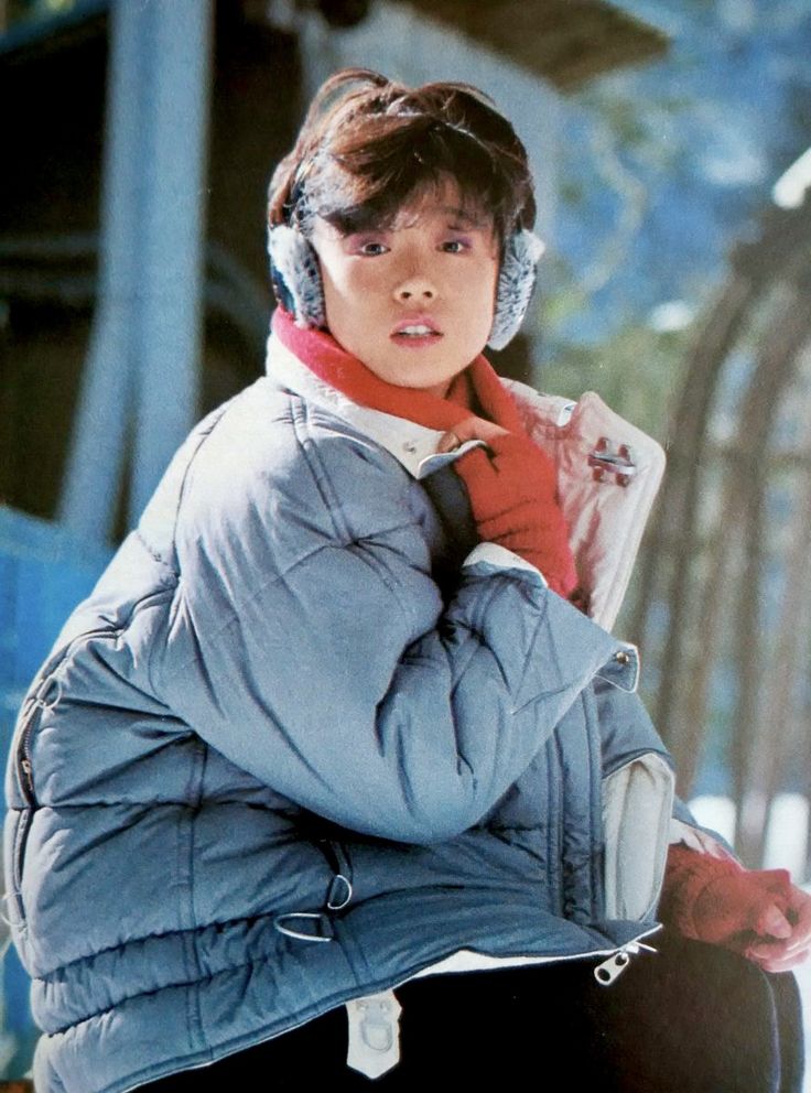 a young girl smiles warmly while wearing a blue winter jacket, a red scarf, and earmuffs.  the image suggests she is enjoying being outdoors in the snow.