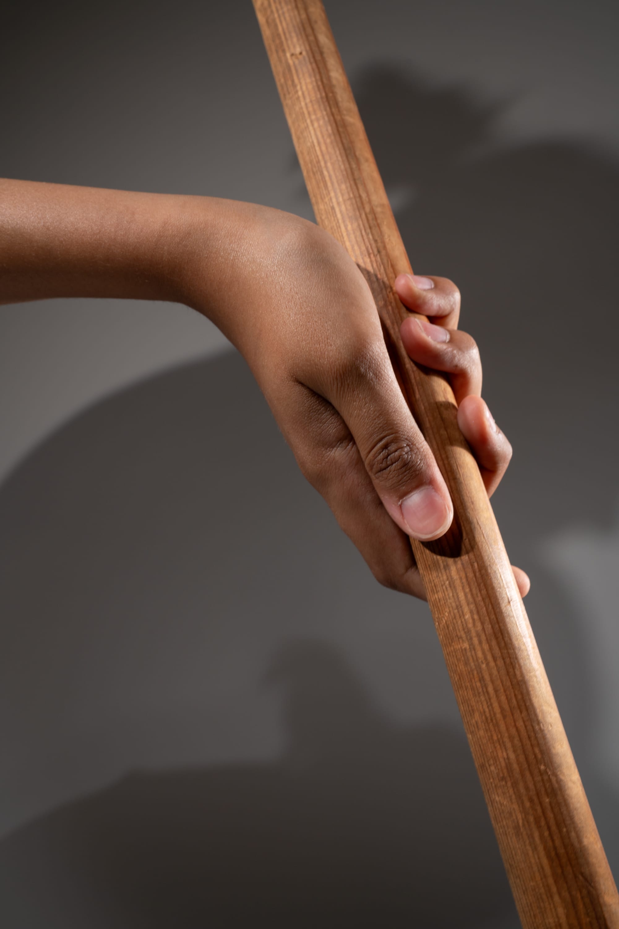 a closeup view of a child's hand tightly gripping a vertical wooden stick. the hand is illuminated by studio lighting, casting a shadow on the wall in the background. the image focuses on the texture and detail of both the hand and the wood, creating a strong sense of tactile awareness.