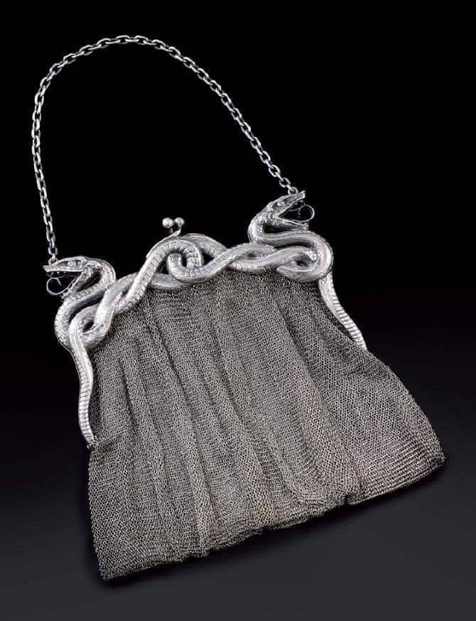the image shows a vintage silver metal mesh purse with a snake design on the frame. the purse has a chain handle and two snake heads on either side of the frame, appearing as though the snakes are holding the chain. the body of the purse is made of tightly woven metal mesh. the background is black.