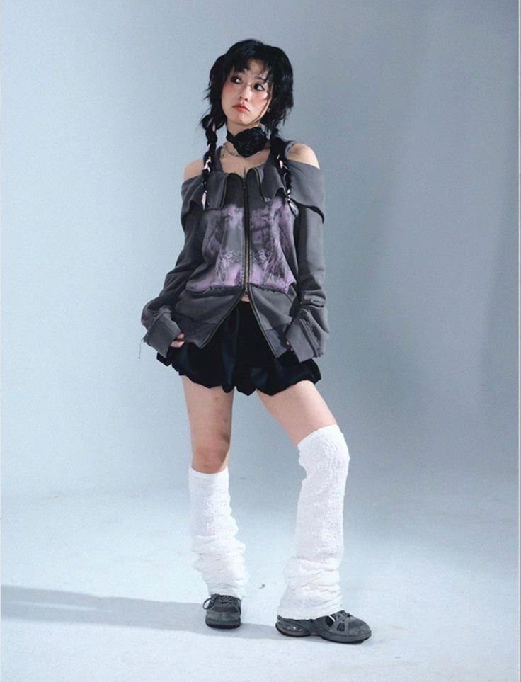 a young woman with dark hair styled in braids is posing in a studio photoshoot. she is wearing a grey offshoulder jacket with purple detailing, a black short skirt, white leg warmers, and dark sneakers. her pose is slightly angled, and she is looking to the side. the background is a light grey, and the overall style is fashionable and edgy.
