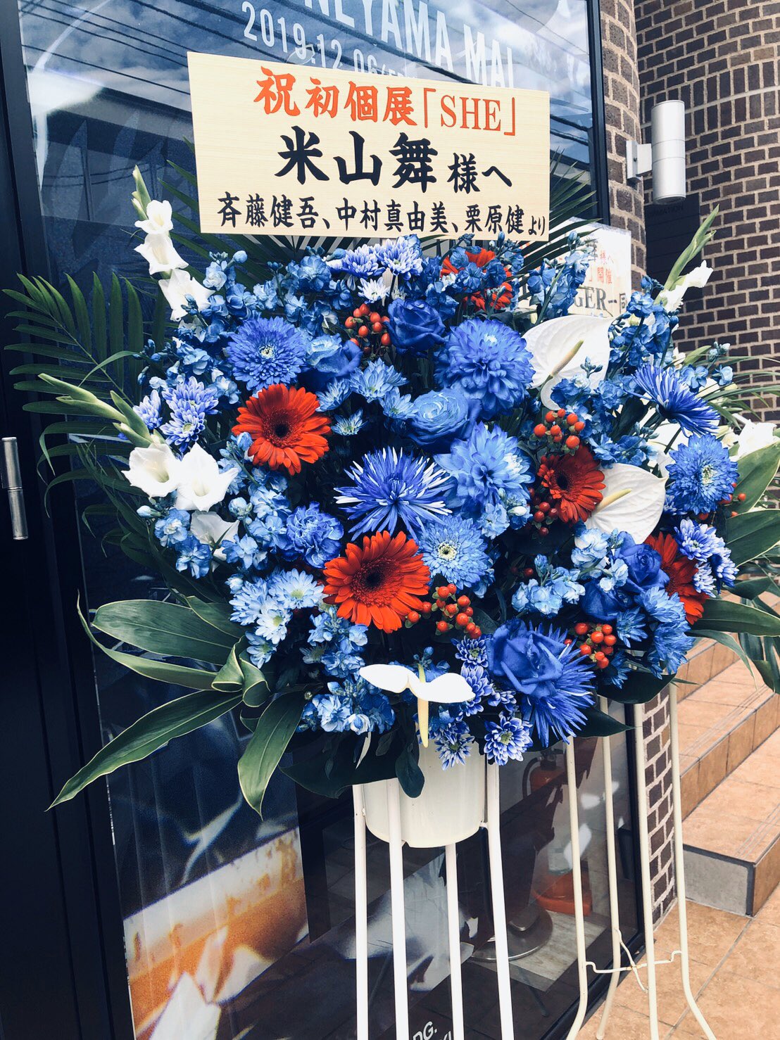 a vibrant flower stand placed in front of a storefront. the stand features an arrangement of red and blue flowers with white accents. a banner on the arrangement congratulates "米山舞" (mai yoneyama) on their first solo exhibition titled "she". it also mentions it's a gift from three people: saito kengo, nakamura mayumi, and kurihara takeshi.