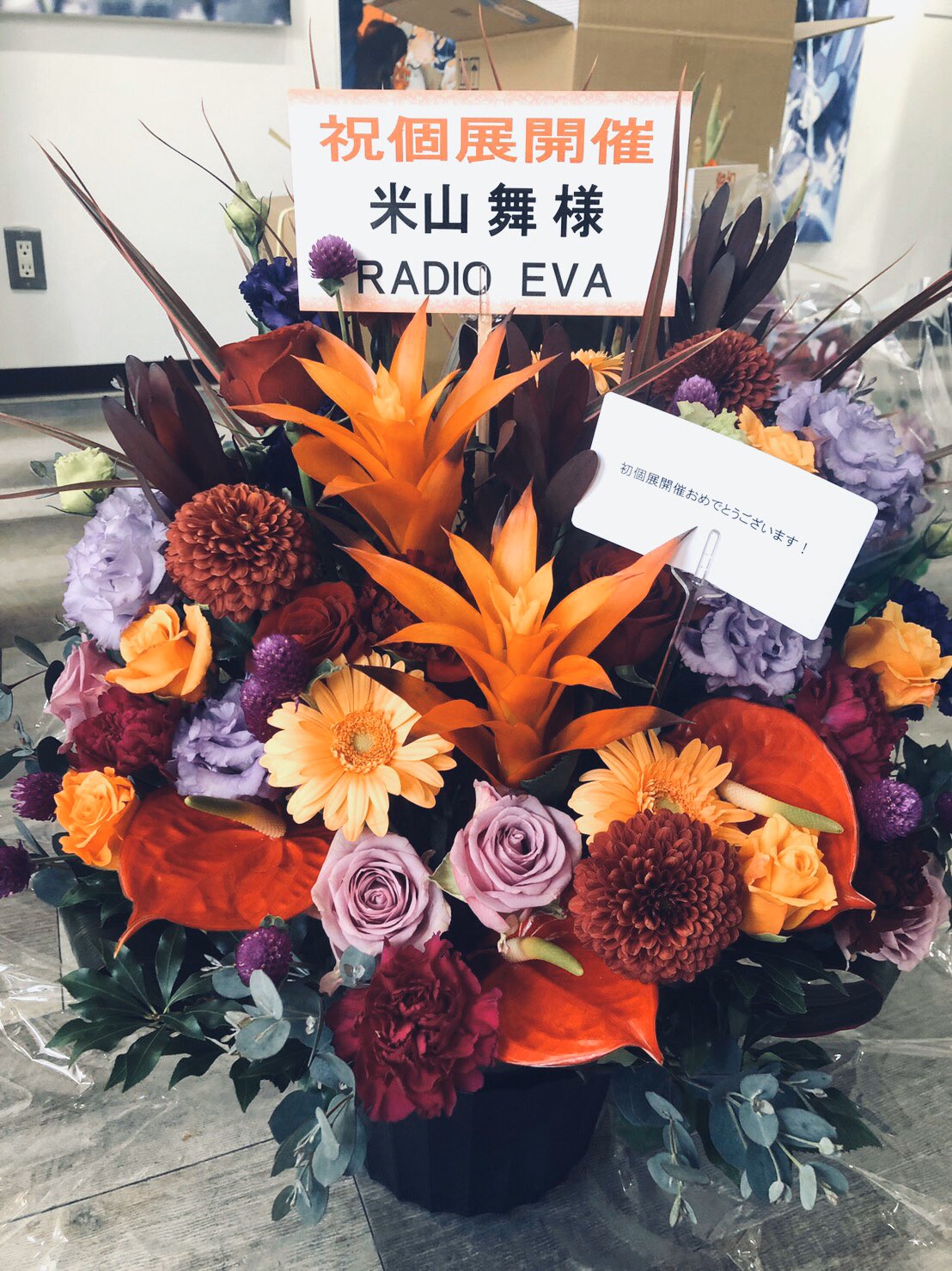 a vibrant flower arrangement with a sign in japanese that appears to be congratulating the opening of something related to "radio eva." the flowers are a mix of orange, red, purple, and yellow.