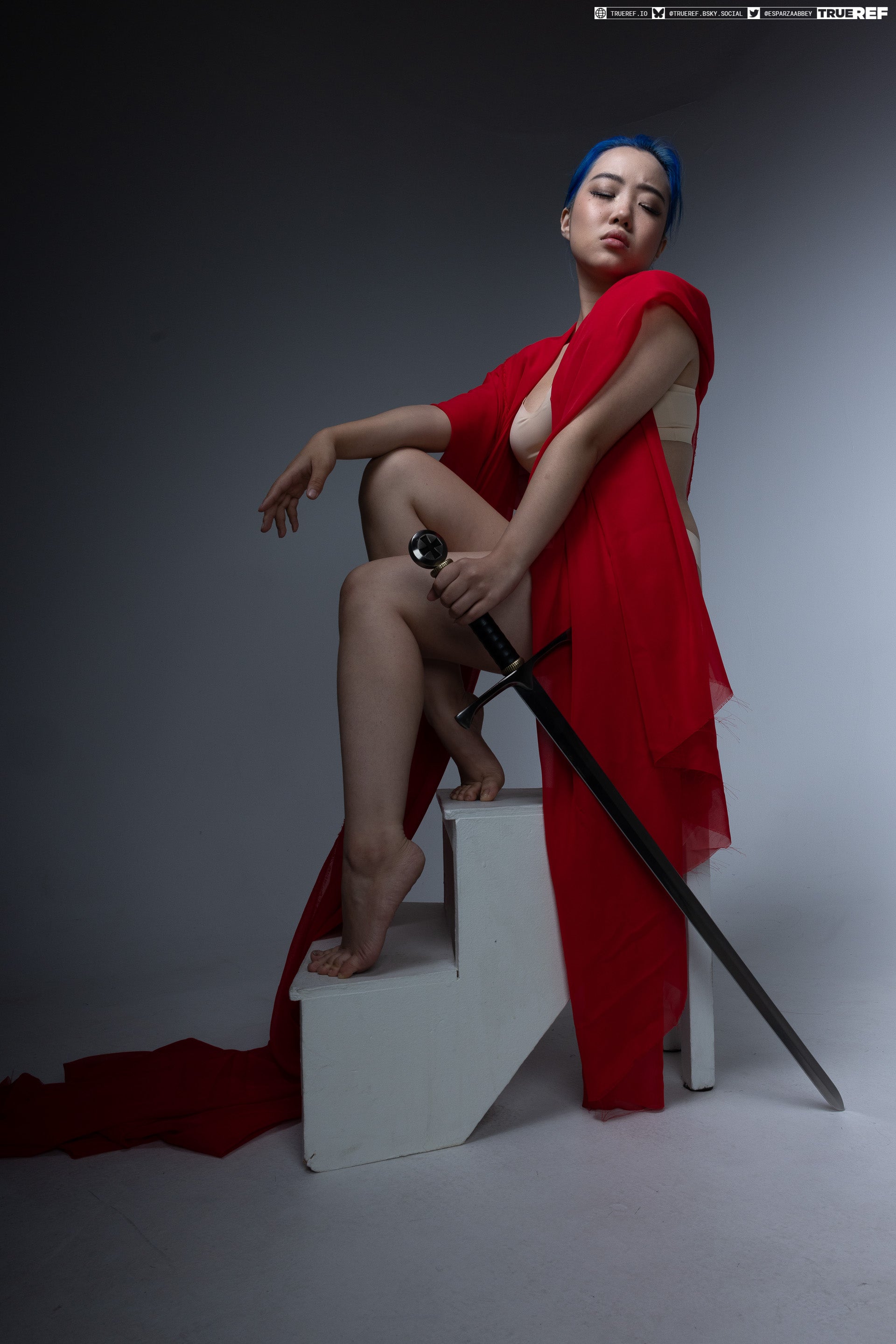 a woman with blue hair is sitting on white stairs. she is wearing a red dress and holding a sword. she is barefoot. the background is a grey studio.