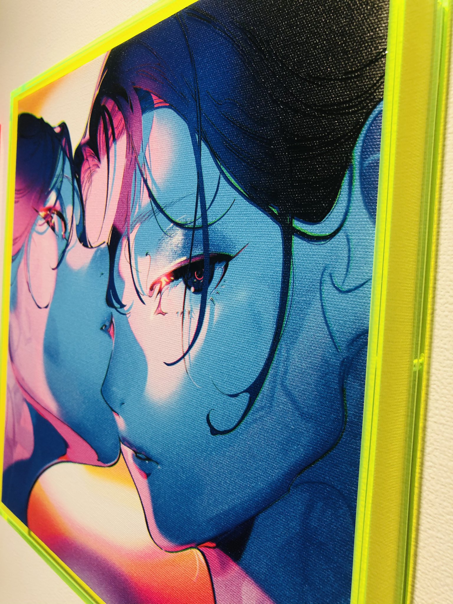 the image shows a detail of a framed digital painting. the artwork is a closeup portrait of two women, their faces rendered in vibrant hues of blue, pink, yellow, and green. the woman in the foreground is partially obscured, her face turned towards the other woman. the background of the painting features a blend of warm and cool colors, creating a sense of depth. the painting is framed in a bright neon yellow frame, which adds to the contemporary and eyecatching nature of the artwork. the setting suggests that the painting is being exhibited in an art gallery.