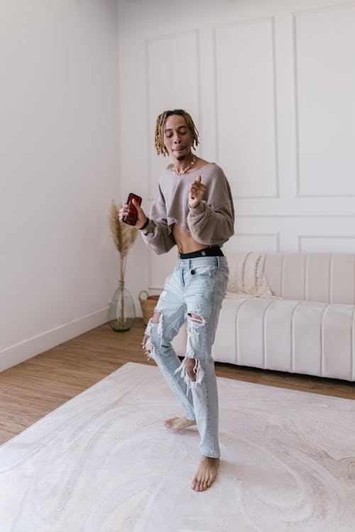 a young man with dreadlocks is joyfully dancing and listening to music from his phone. he's wearing a beige cropped sweater, light wash ripped jeans and is barefoot.  he is in a bright, minimalist living room with a white couch and rug. the overall vibe is carefree and stylish.