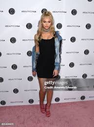 a woman stands on a pink carpet in front of a beautycon backdrop. she is wearing a black dress, a denim jacket, and red high heels. she has blonde hair pulled back in a ponytail and is wearing a choker necklace.