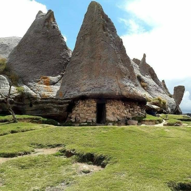 the image depicts a stone house nestled within unique rock formations. the house appears to be built using stones, with a dark entrance. above it, a large, peaked rock formation acts as a natural roof. the surrounding landscape is covered with green grass, suggesting a rural or mountainous setting. the sky is visible in the background with some clouds, indicating it's daytime. other rock formations are visible in the background.