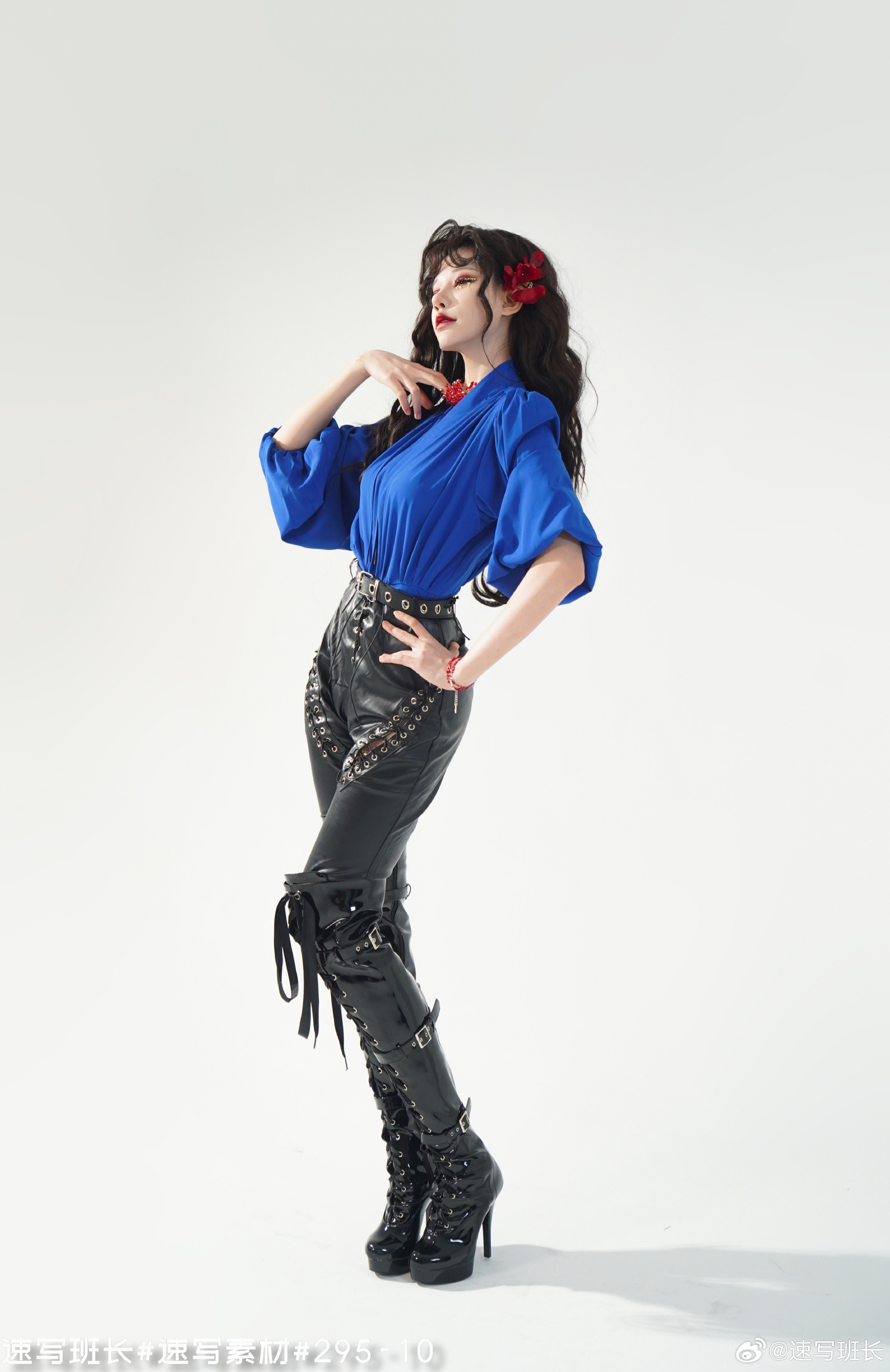 a woman with long black curly hair and a red flower tucked behind her ear stands in a bright studio. she is wearing a blue long sleeve shirt, black leather pants with buckles, and knee high black leather high heel boots. she is accessorized with red bracelets on both wrists and a red choker necklace. the woman is striking a pose with her head tilted back and her hands resting on her chest. the image contains chinese characters at the bottom left.