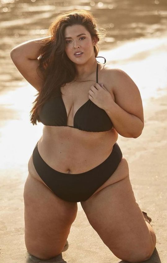 a woman is kneeling on a beach wearing a black bikini. she has her hand on her hair and is looking at the camera. the background is a beach with the ocean. the woman is plussize with brown hair.