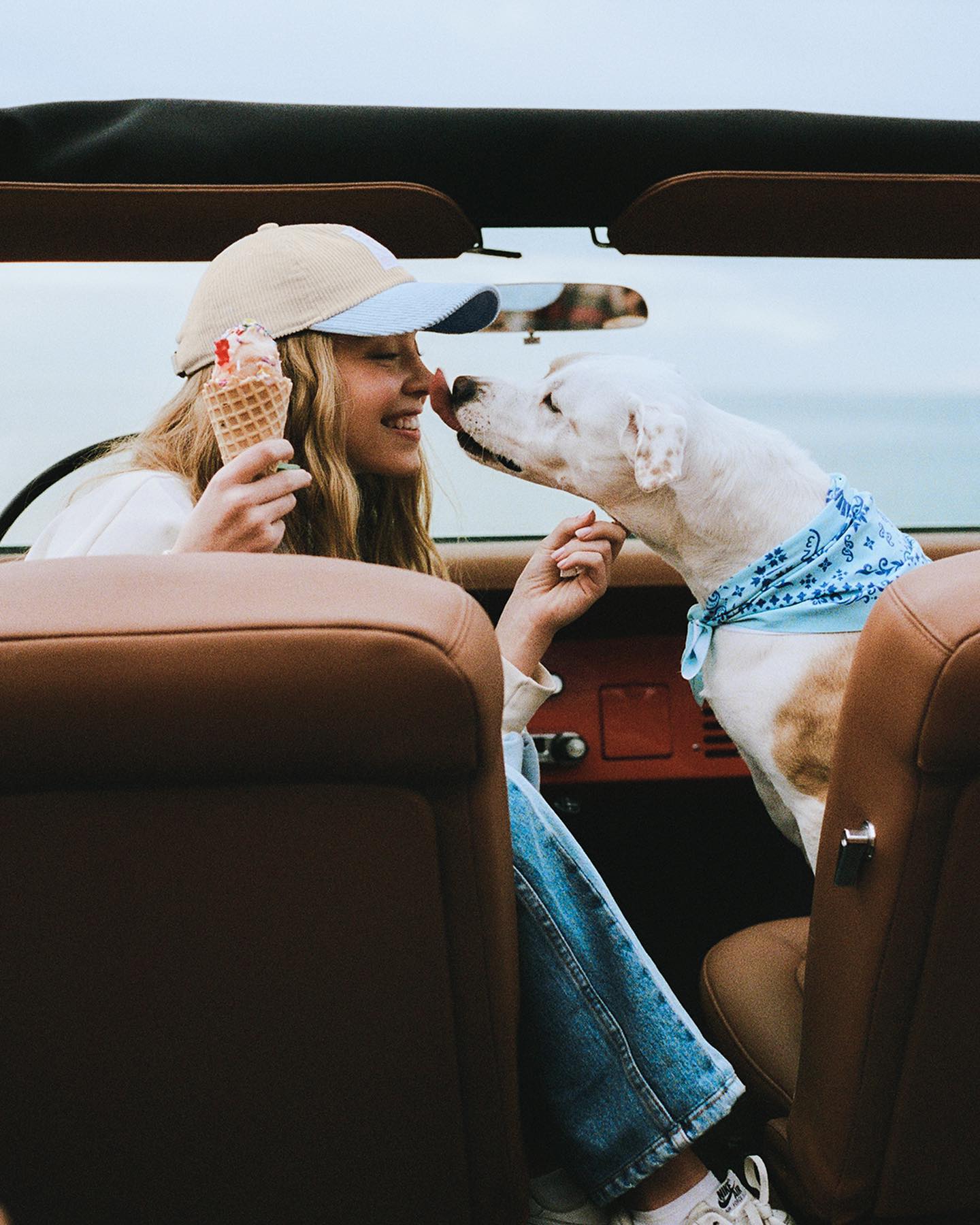 a young woman wearing a beige baseball cap and jeans sits in the front seat of a convertible car with her dog. she is holding an ice cream cone and sharing it with her dog, who is wearing a blue bandana. they appear to be on a road trip and are enjoying their time together. the scene is bright and sunny, and the overall mood is happy and carefree.