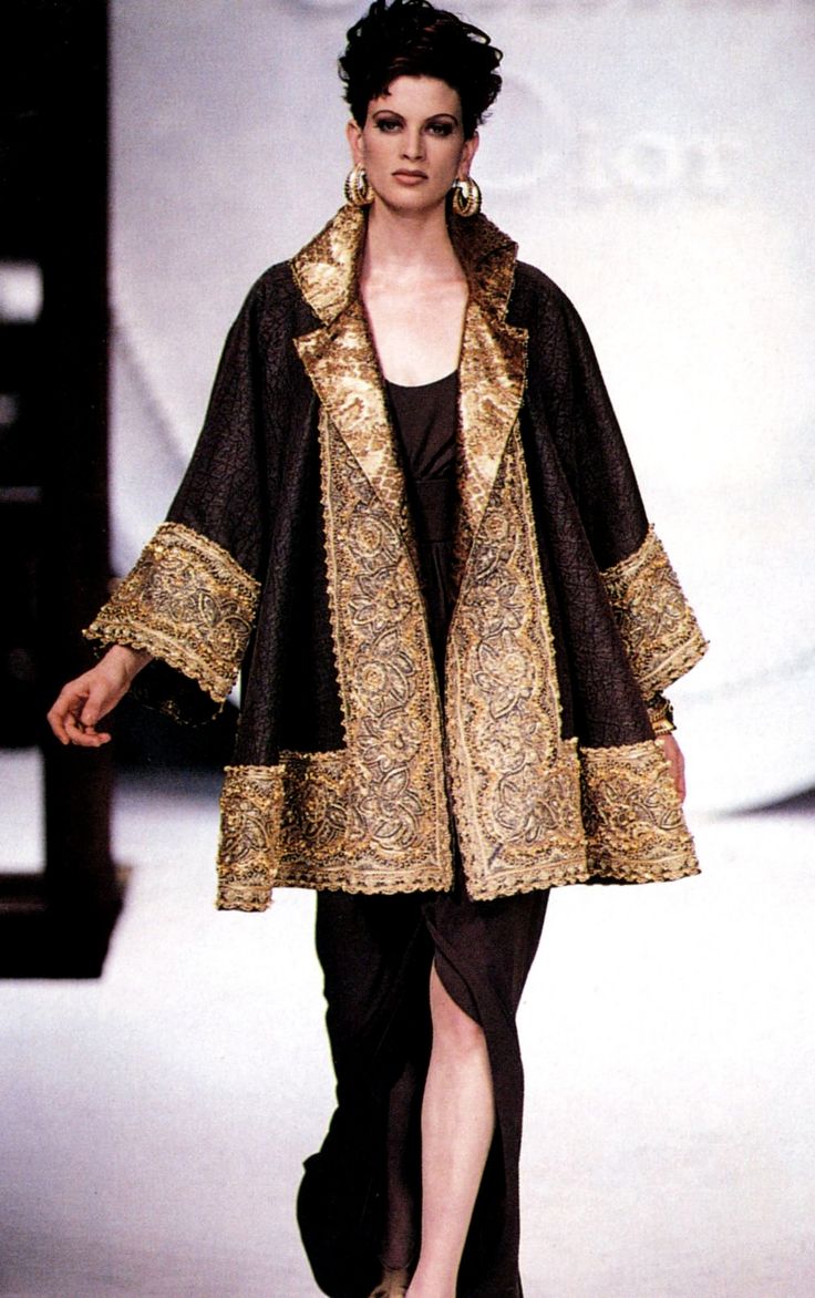 the image shows a fashion model walking a runway. she is wearing a striking dark brown coat or jacket with elaborate gold embroidery detailing on the collar, cuffs, and hem. the coat is kneelength, features wide sleeves, and is worn over a dark brown, floorlength dress with a high slit. the model's hairstyle is a short, structured style, and she wears large gold hoop earrings. the backdrop is a minimalist, bright runway setting.  the overall style evokes a 1990s high fashion aesthetic.