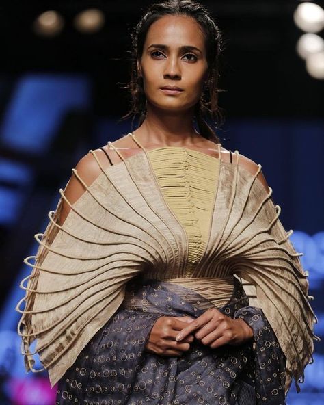 a model poses on a runway showcasing a unique fashion design. she wears an intricate dress with a layered, pleated upper portion in a beige or light gold color, featuring structural elements that radiate from the chest. the lower part of the dress is a patterned, darkertoned fabric wrapped around her waist. the stage is dimly lit with a dark blue backdrop. the model has a serious expression and her hair is neatly styled.