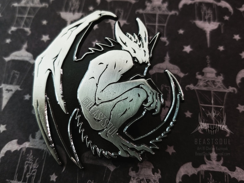 loseup shot of a silver and black enamel pin featuring a gargoylelike creature with wings. the creature is depicted in a crouching position, its body mostly silver with black accents. the pin is set against a dark background featuring a repeating pattern of lanterns and stars. the artist's logo and contact information are visible in the bottom right corner. the pin's design is highly detailed and intricate. the overall aesthetic is gothic and dark fantasy.