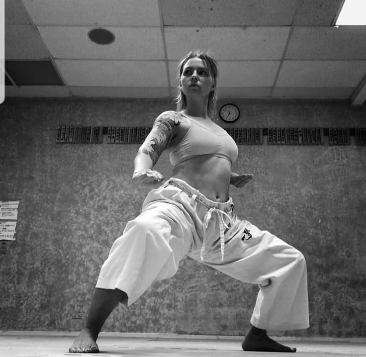 a woman is in a martial arts stance, likely karate, wearing workout clothes and displaying a determined expression. she has a tattoo on her arm. the photo is in black and white, taken in a gymnasium setting with a clock visible on the wall. she is barefoot and appears to be training or performing a kata.