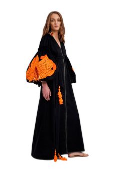 a woman wearing a long black dress with orange floral embroidery, standing in front of a white background. she is looking off to the side, with a relaxed yet elegant expression. the dress has long sleeves, and is fitted at the waist, with a flowing skirt. the embroidery is delicate and intricate, adding a touch of femininity to the dress. the woman's hair is long and flowing, framing her face beautifully. the dress is a perfect choice for a summer occasion, and is sure to turn heads.