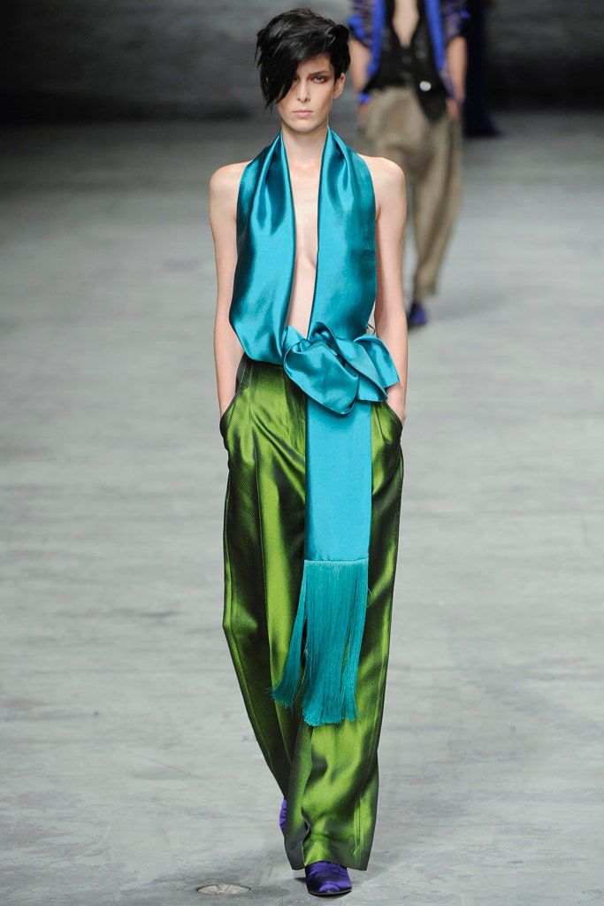 a model in a green satin jumpsuit and a long blue scarf walks the runway. she is wearing a blue and green outfit, with a green satin jumpsuit and a long blue scarf. she has her hair styled in a short, choppy cut, and she is walking with a confident stride. the background is blurred, and there is a man in the background in a blue shirt and brown pants.