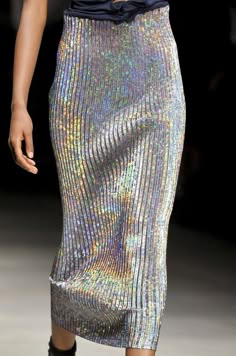 a model walks down the runway in a silver sequined skirt. she is posing with her legs crossed and her arms held at her sides. the skirt is very sparkly and reflects the light. the model is wearing a black top, which is not visible in the image.