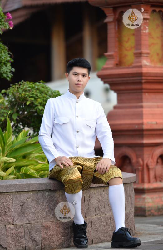 a young thai man wearing traditional white shirt with gold patterned shorts and white socks and black shoes, sitting on a stone wall in front of a temple, smiling and looking at the camera.