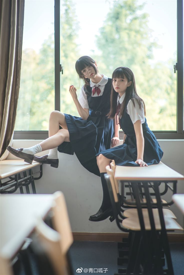 two young women in school uniforms are sitting on desks in a classroom. the first woman is sitting on the desk by the window. she has her legs crossed and is wearing black loafers and white socks. the second woman is sitting on the desk to the right of the first woman. she is wearing black loafers and black stockings. both women are wearing white shirts and dark blue jumpers with red bows.