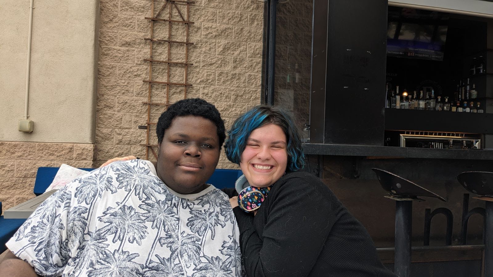 two friends, a man and a woman, are sitting outside a restaurant with their arms around each other. they are both smiling at the camera. the man is wearing a white shirt with black flowers on it, and the woman is wearing a black shirt. the restaurant has a black bar and black stools. the image is taken from eye level and the lighting is natural and daytime.