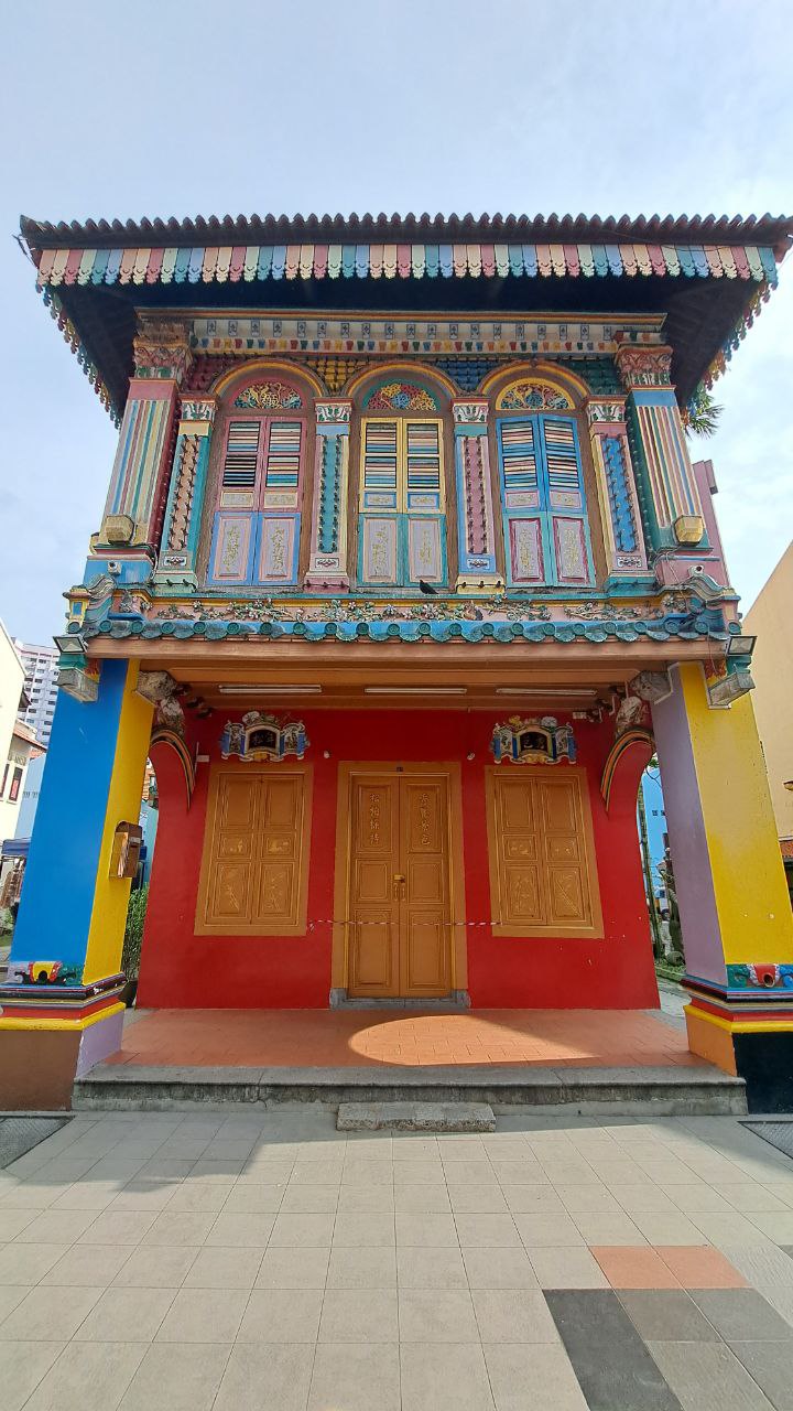 a brightly colored shophouse in singapore. the facade of the building is decorated with intricate details and features a mix of colors, including red, blue, yellow, teal, and brown. the windows and doors are all adorned with ornate carvings. the building is located on a street with a concrete sidewalk. the photograph captures the vibrant and unique architecture of singapore.