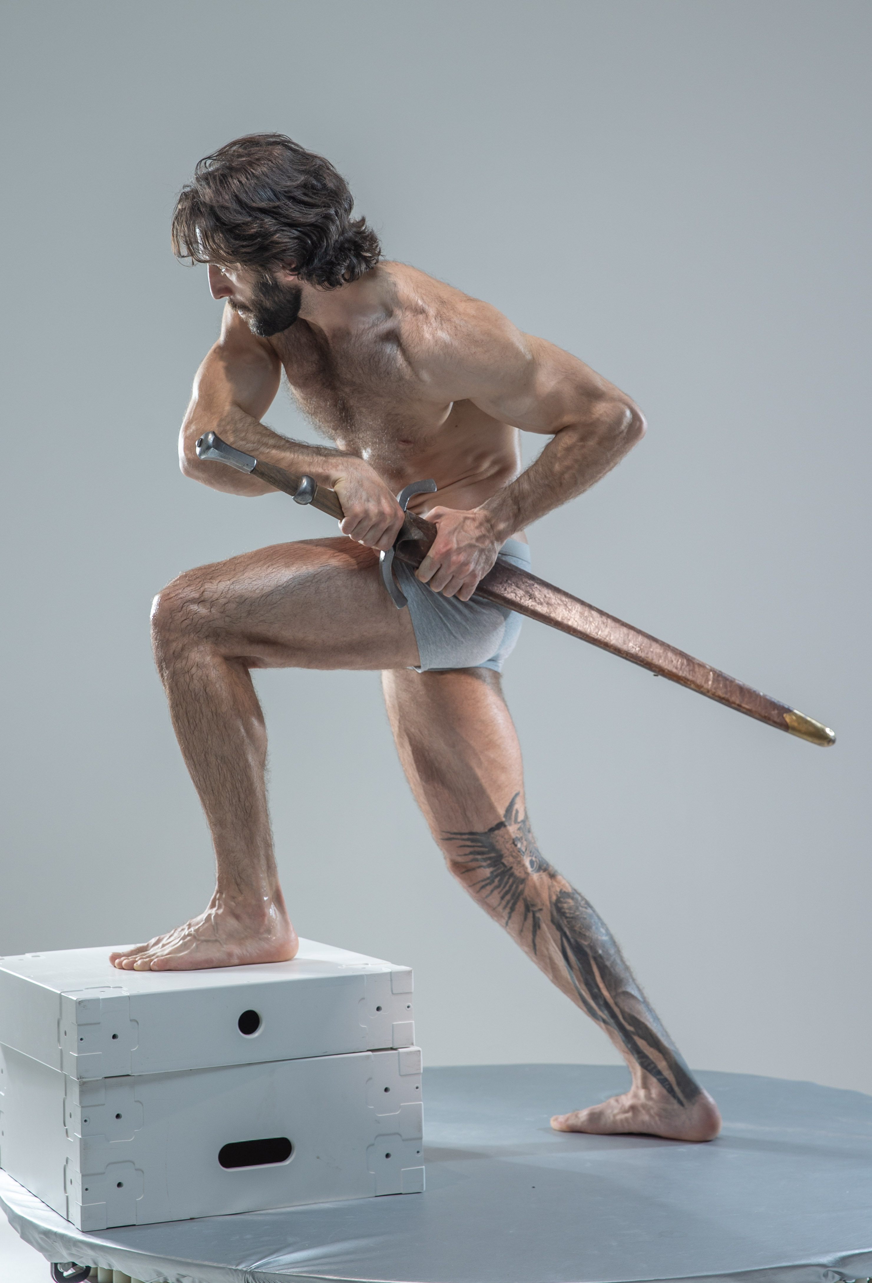 a muscular man with long hair and beard stands barefoot on a white platform in a fighting stance. he holds a long sword with both hands, his body tensed in preparation for combat.  he is shirtless, wearing only gray underwear, showcasing his physique. his left leg is extended back for balance, revealing a large tattoo of a bird on his calf. the background is a neutral gray, indicative of a studio setting.  the lighting is bright and artificial, highlighting the details of his muscles and the sword.