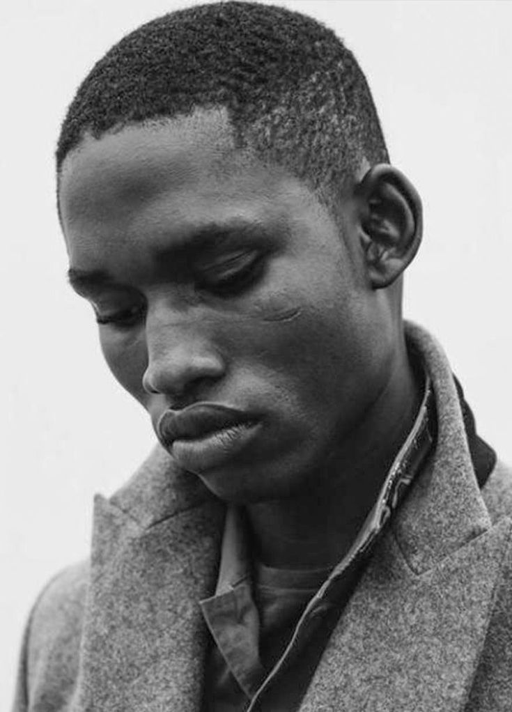 a black and white closeup portrait of a seriouslooking man with short hair. he is wearing a grey textured coat and has a thoughtful expression on his face.  his skin is dark, and a small scar is visible on his cheek. the image focuses on his face and upper body, emphasizing the textures of his skin and clothing.  the overall mood is contemplative and stylish.