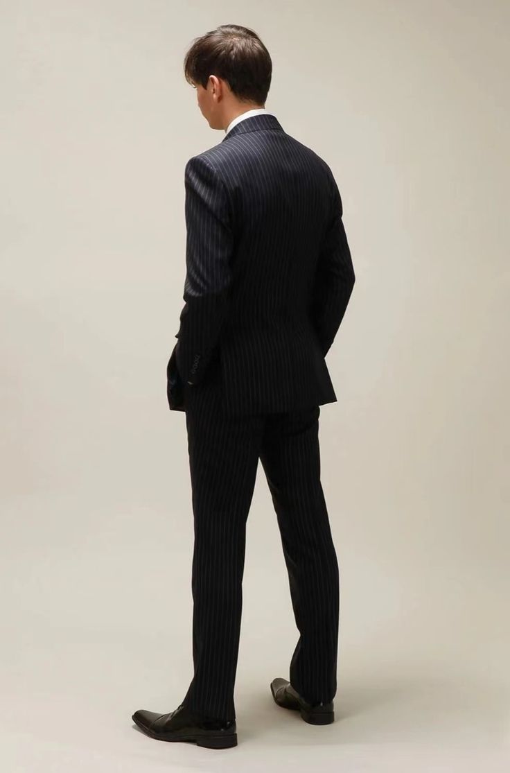 a man in a dark navy blue pinstripe suit stands with his back turned to the camera, looking over his shoulder. the suit is welltailored and fits him perfectly. he is wearing black shoes and has a cleancut look. the background is a plain light gray. the overall image conveys a sense of professionalism and confidence.