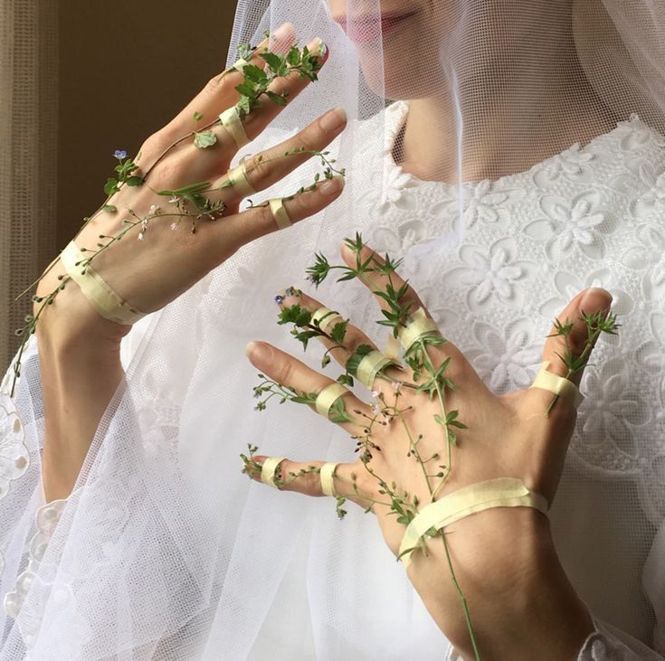 the image showcases a closeup view of a bride's hands adorned with small sprigs of wildflowers and greenery delicately secured with pale yellow tape.  the bride is wearing a white, lacy dress and a sheer veil that drapes over her.  the overall aesthetic is very soft, romantic, and ethereal, emphasizing the delicate beauty of the flowers and the bride's hands. the image appears to be a carefully styled photograph, possibly for artistic purposes or as part of a wedding shoot. the focus is on the unique floral arrangement on the hands, creating a whimsical and natural contrast with the traditional wedding attire.