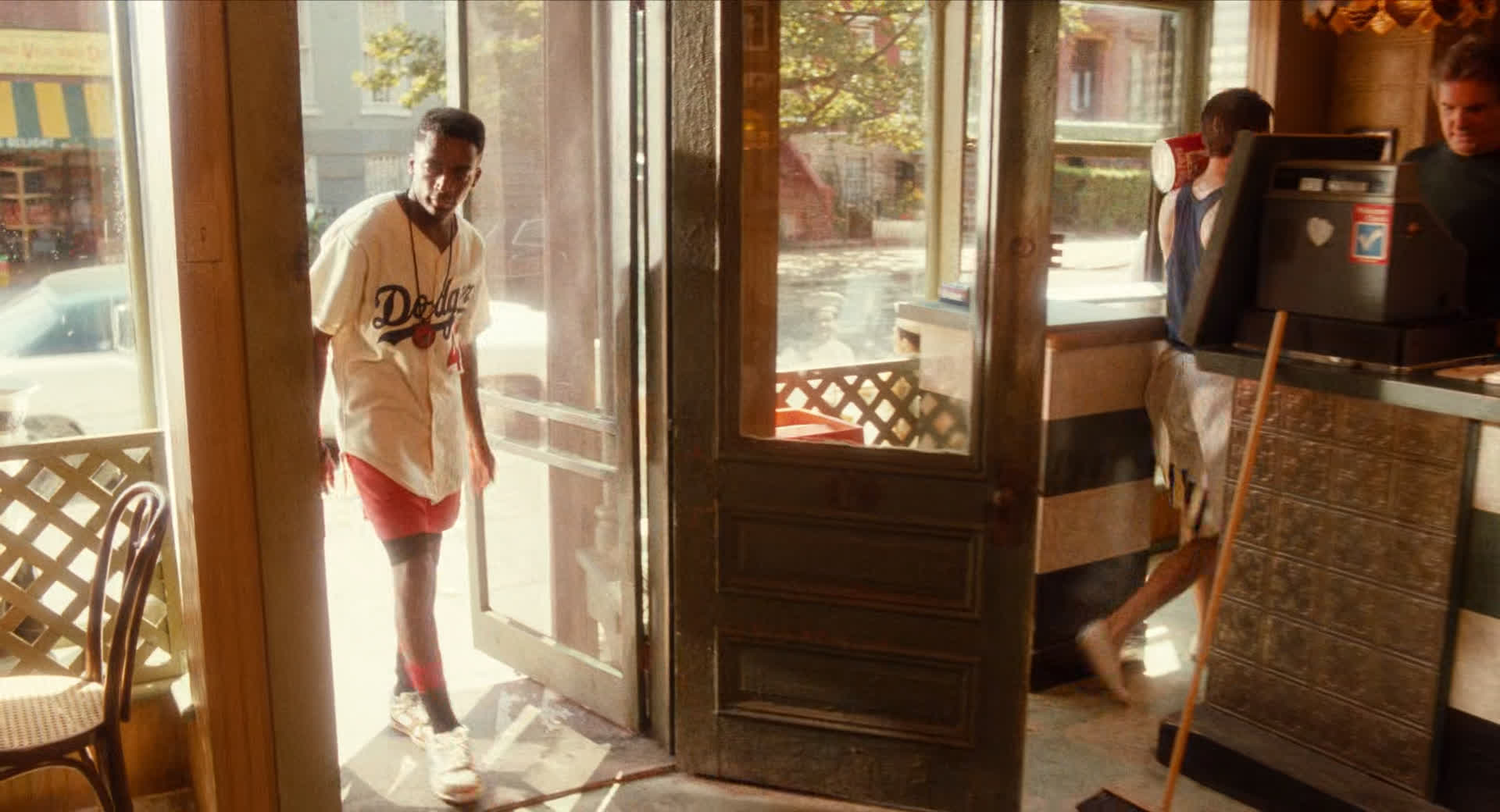 a man wearing a baseball jersey is walking into a restaurant. the interior of the restaurant is dark and cool, while the street outside is bright and sunny. the door is open, and we can see the street and a few cars through the doorway. the man is wearing red shorts and white sneakers. the restaurant has a green and white checkered floor. there are two other people in the restaurant, one of whom is working behind the counter.