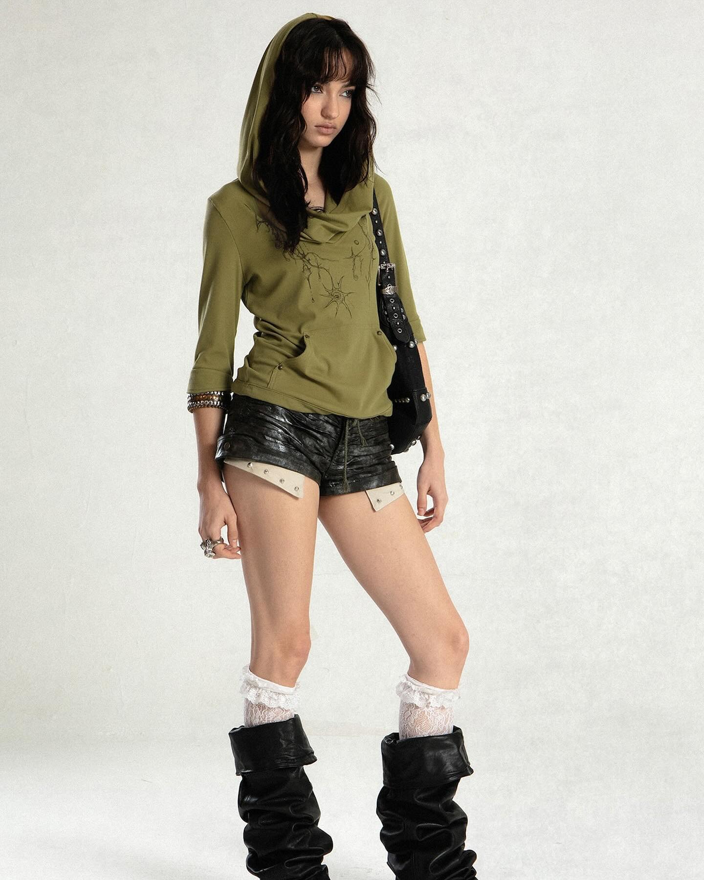a young woman with long brown hair is standing in a studio wearing a stylish outfit. she has on an olive green hooded sweatshirt, black leather shorts with beige detailing, white lace socks, and slouchy black boots.  she carries a black handbag over her shoulder. the overall style is edgy and alternative.  the background is a plain light beige.