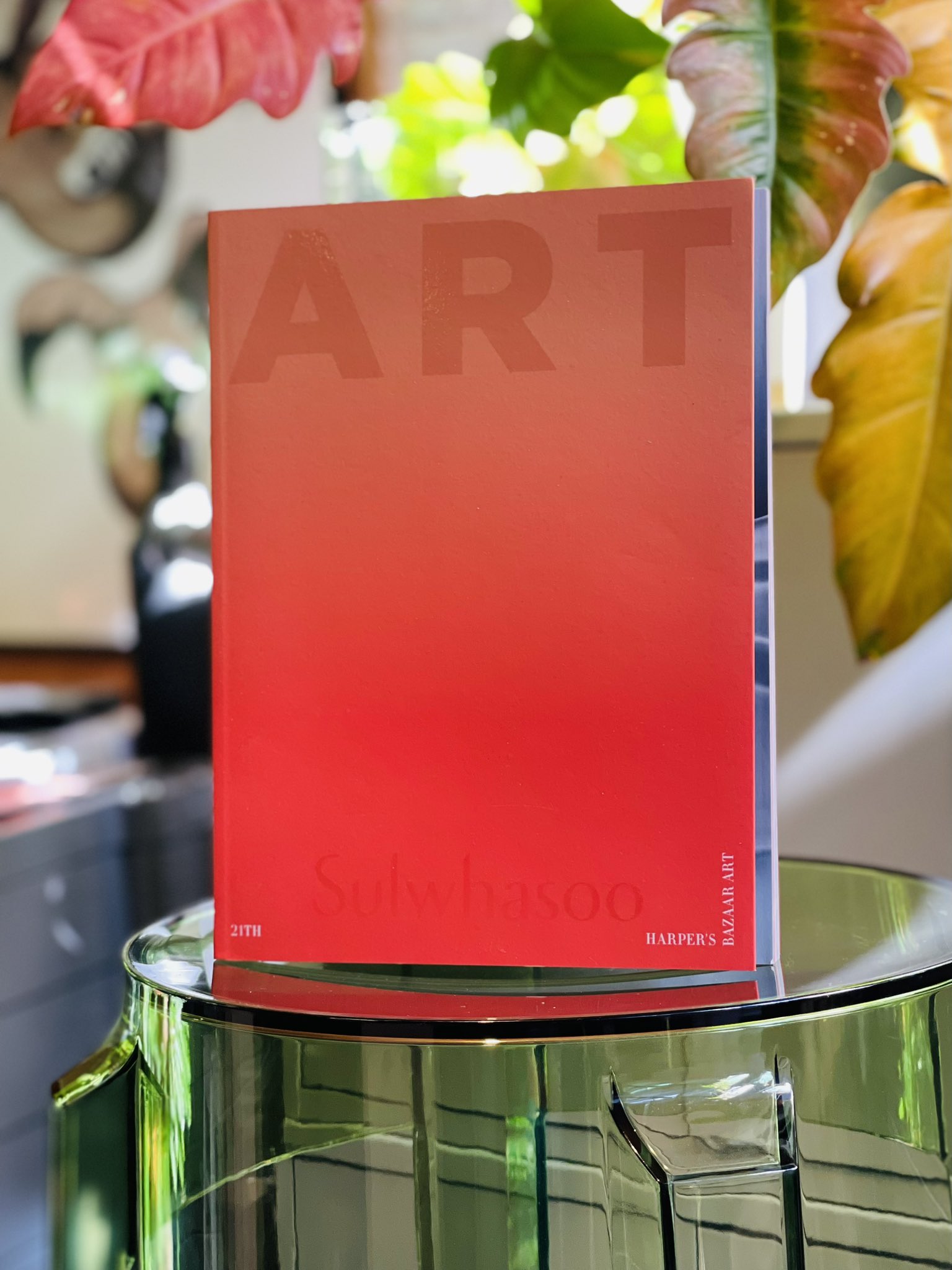 a red art book with a gradient from light pink to red, titled "art" and "harper's bazaar art" is standing upright on a clear glass table. there is a green house plant behind the table, and artwork visible in the background.