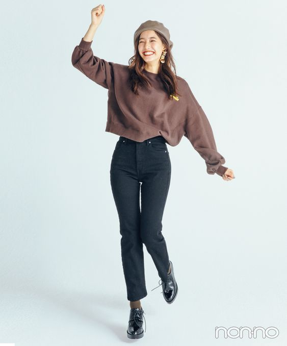 a woman is standing in front of a white background, posing for the camera. she is wearing a brown sweatshirt, black jeans, a beige beret, and black shoes. she is also wearing a pair of earrings. she is smiling and appears to be happy.