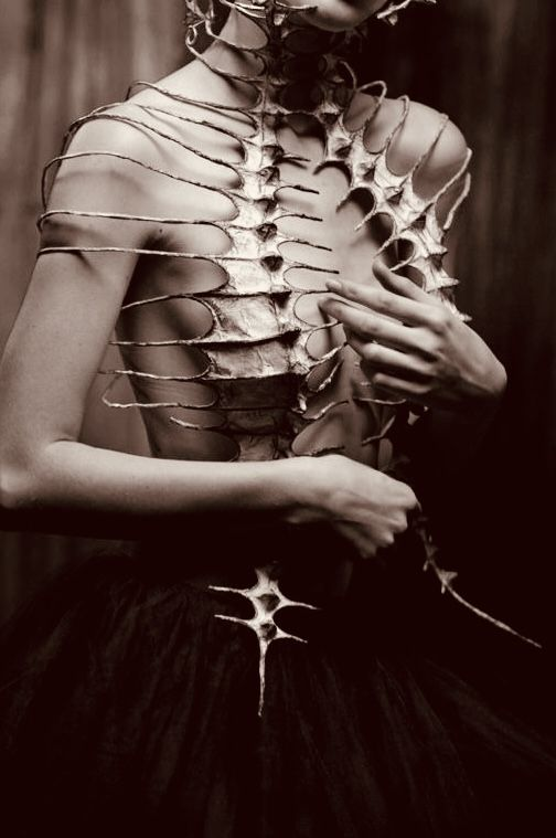 a black and white image of a slim woman, possibly a model, wearing an elaborate skeletallike bodice made of a light material. the bodice is intricate with delicate, spiky protrusions that resemble thorns or bones. it covers her torso and extends to a chokerlike piece around her neck. she wears a dark, possibly black, tutulike skirt that contrasts with the bodice. her pose is elegant, with her hands resting gently on her stomach, drawing attention to the unique garment. the image is likely a stylized fashion photograph or artistic representation, emphasizing the artistry of the costume and the model's pose.