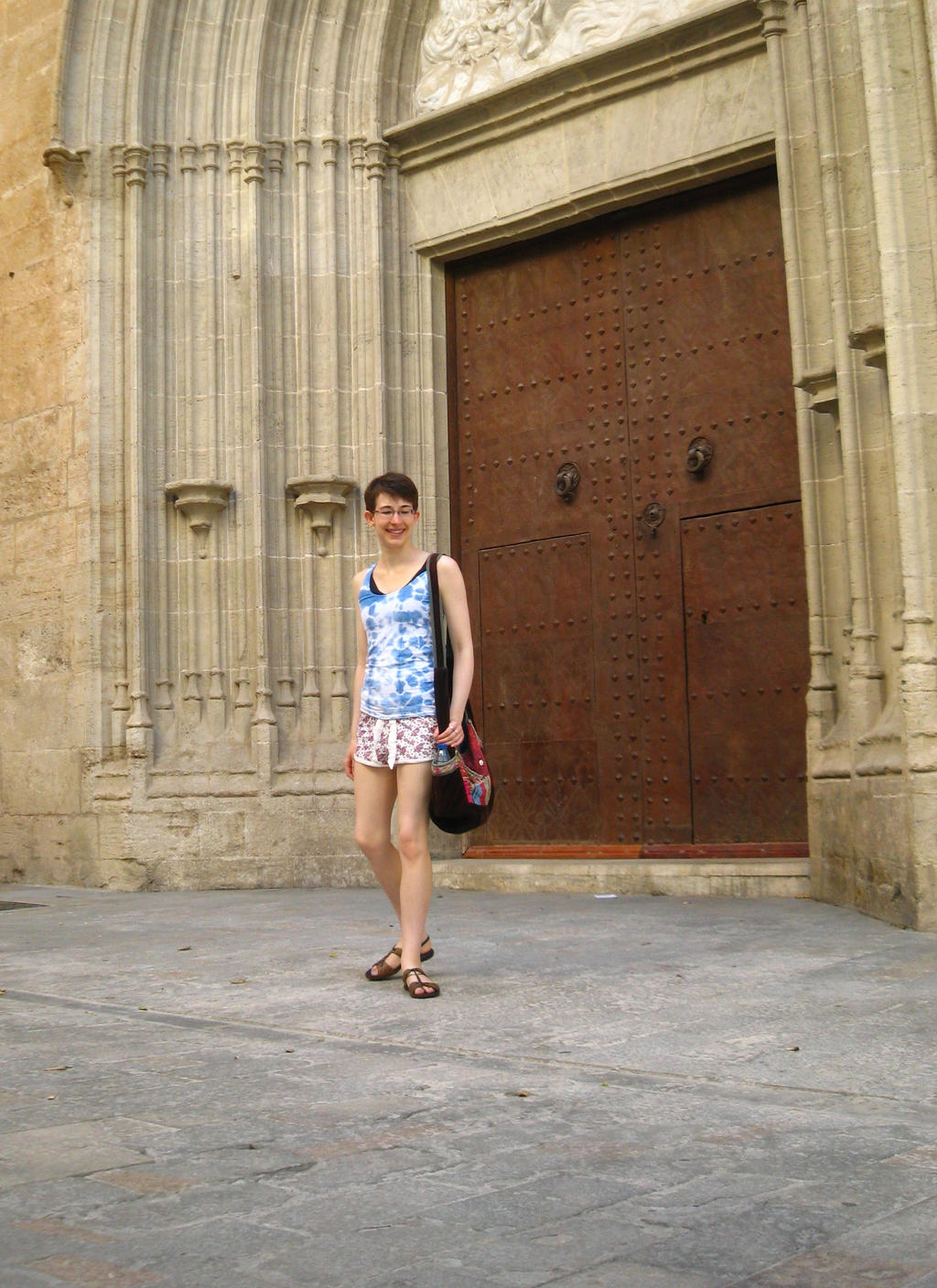 a young woman stands in front of a large, old wooden door in a stone building. she is wearing a blue and white floral tank top and shorts, sandals, and carrying a brown purse. she is smiling and looking at the camera. the architecture is typical of a spanish or european old town.