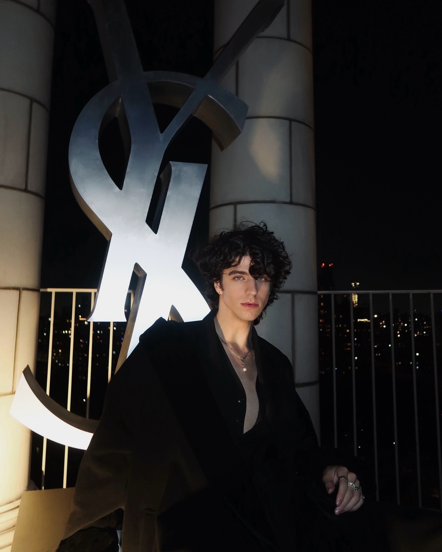 a portrait of a man with curly hair wearing a black coat and jewelry. he is sitting in front of a large silver ysl logo on a rooftop at night. city lights are visible in the background behind a railing.