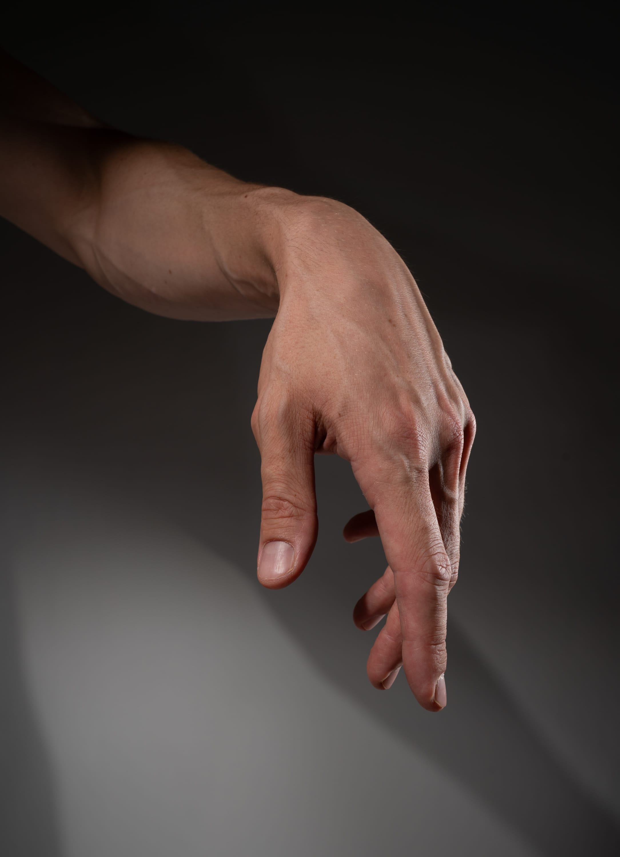 the image showcases a man's right arm and hand extended downward against a gray background. the hand is slightly open with fingers relaxed, creating a natural and effortless gesture. the lighting is dramatic, with a spotlight highlighting the arm and hand while casting shadows on the background. this creates a sense of depth and emphasizes the contours of the subject.
