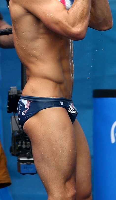 a closeup of a muscular male swimmer in a blue swimsuit, showing off his torso, abs, and wet skin. the swimmer appears to be in competition and is focused and ready to perform. he is wet from swimming and the water droplets are visible on his skin. he is wearing a blue swimsuit with a white adidas logo.