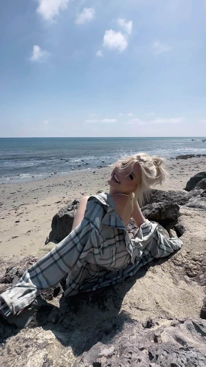 a woman with blonde hair, wearing a plaid shirt, is sitting on rocks at the beach, smiling and looking up at the sky. the ocean is visible in the background under a sunny sky with some clouds.