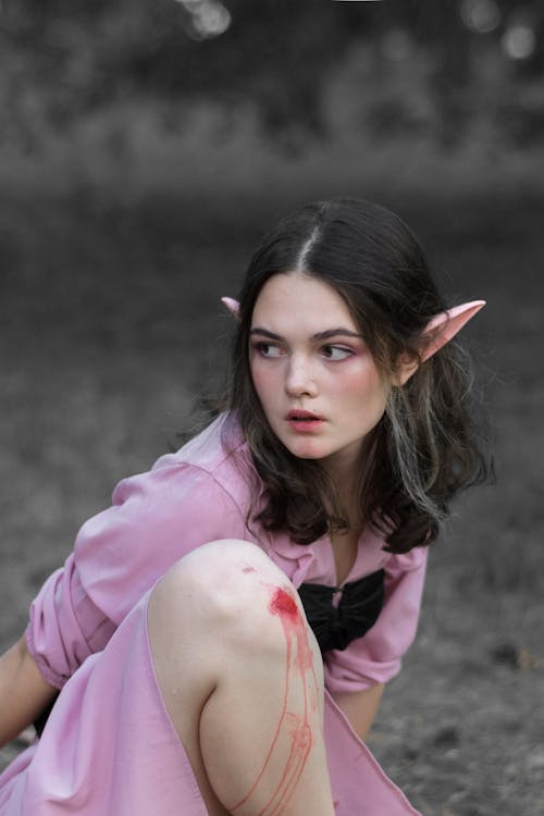 a young woman with elf ears is outdoors. she is wearing a pink shirt and has a scrape on her knee with blood running down her leg. she is looking away from the camera. the background is a blurred grey.