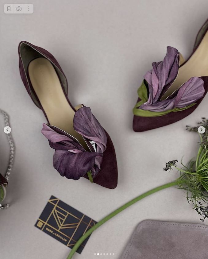 the image showcases a pair of stylish shoes adorned with flower embellishments, presented in a flat lay composition. the shoes are crafted from suede material and feature a deep purple hue. delicate purple flowers accent the front of the shoes, adding an artistic and decorative touch. the presentation also includes a business card, sprigs of lavender, and other accessories, contributing to an overall aesthetic of elegance and modern style.