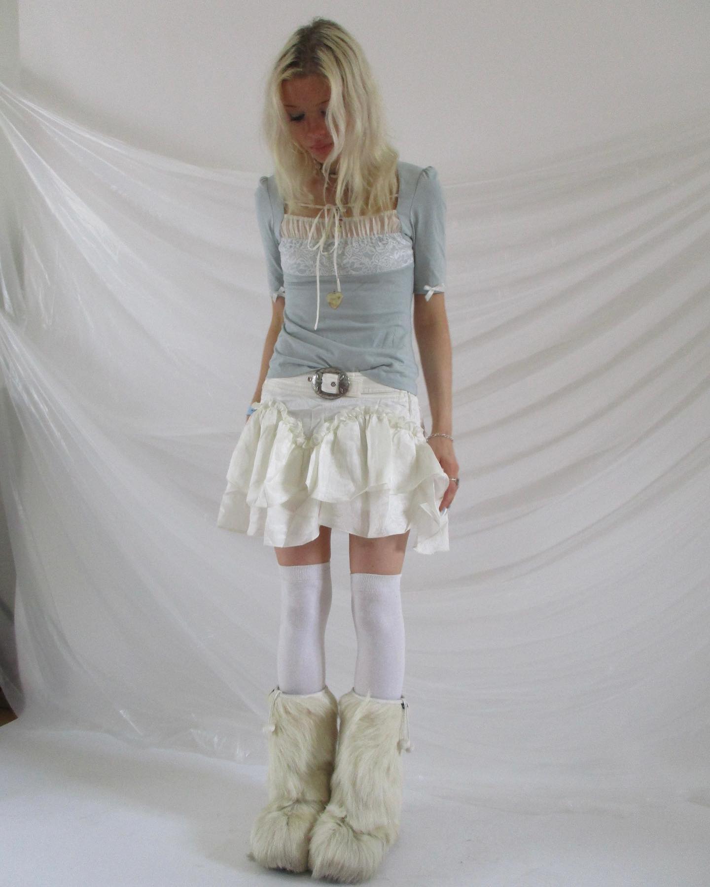 a young woman stands in a studio shot against a white backdrop, showcasing a charming outfit. she wears a light blue, shortsleeved top with lace detailing at the neckline and a ruffled white skirt.  her accessories include a white belt and a delicate necklace.  she is also wearing white kneehigh socks and fluffy white boots that extend to her midcalf. the overall aesthetic is ethereal, suggesting a blend of innocent and whimsical fashion.  her blonde hair is styled simply, framing her face. the image emphasizes the texture of the clothing and the softness of the white background.