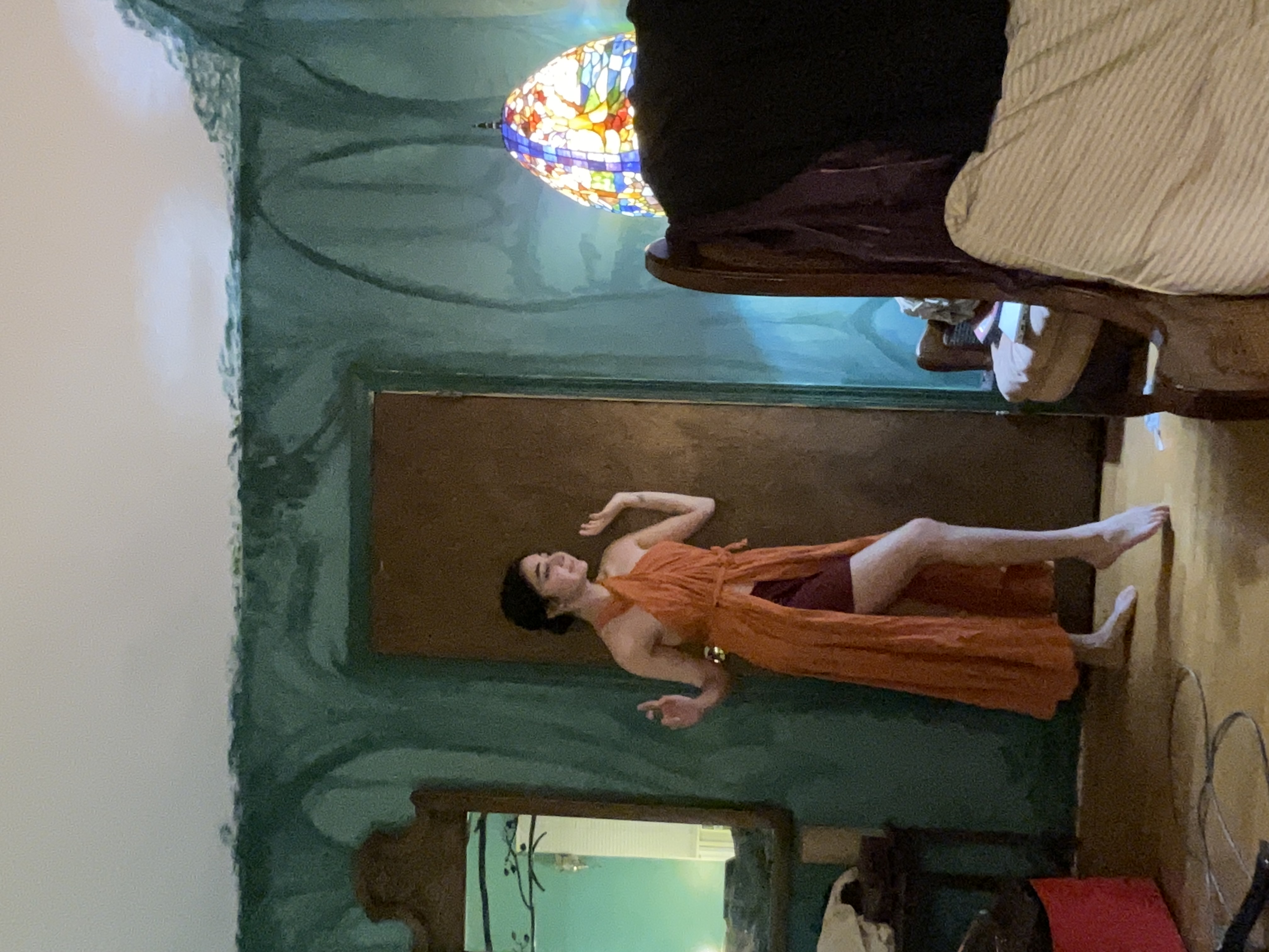 a young woman stands in a bedroom with a uniquely painted green wall. she is wearing a long, orange halter dress with a high slit. the room features antique furniture, including a chair and a bed, and a stained glass lamp. the woman is posing for a photo.