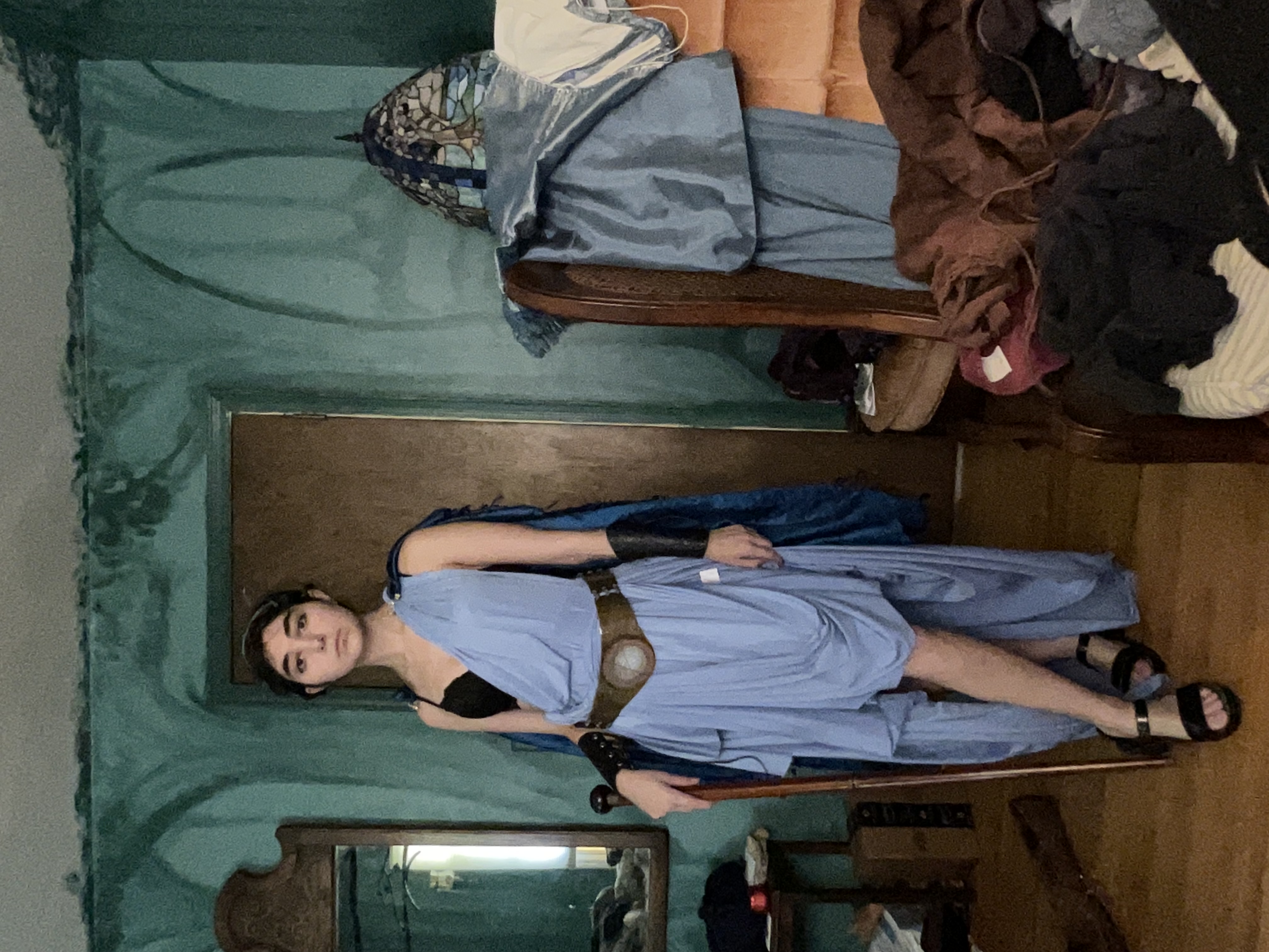 a young woman is posing for a photograph in a homemade cosplay costume. she is dressed as a greek goddess, wearing a light blue draped dress and sandals. she holds a wooden staff in her left hand. the background features a messy room with teal walls, a chair piled with clothes and blankets, and a wooden chair. the overall mood is casual and informal.