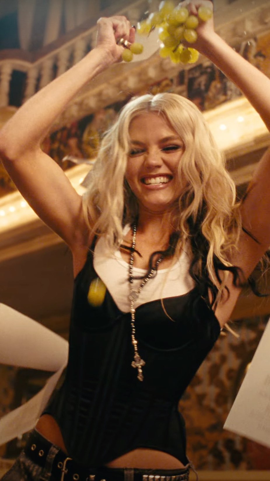 a blonde woman wearing a black corset and a necklace is smiling while holding a bunch of grapes above her head with her arms raised indoors.